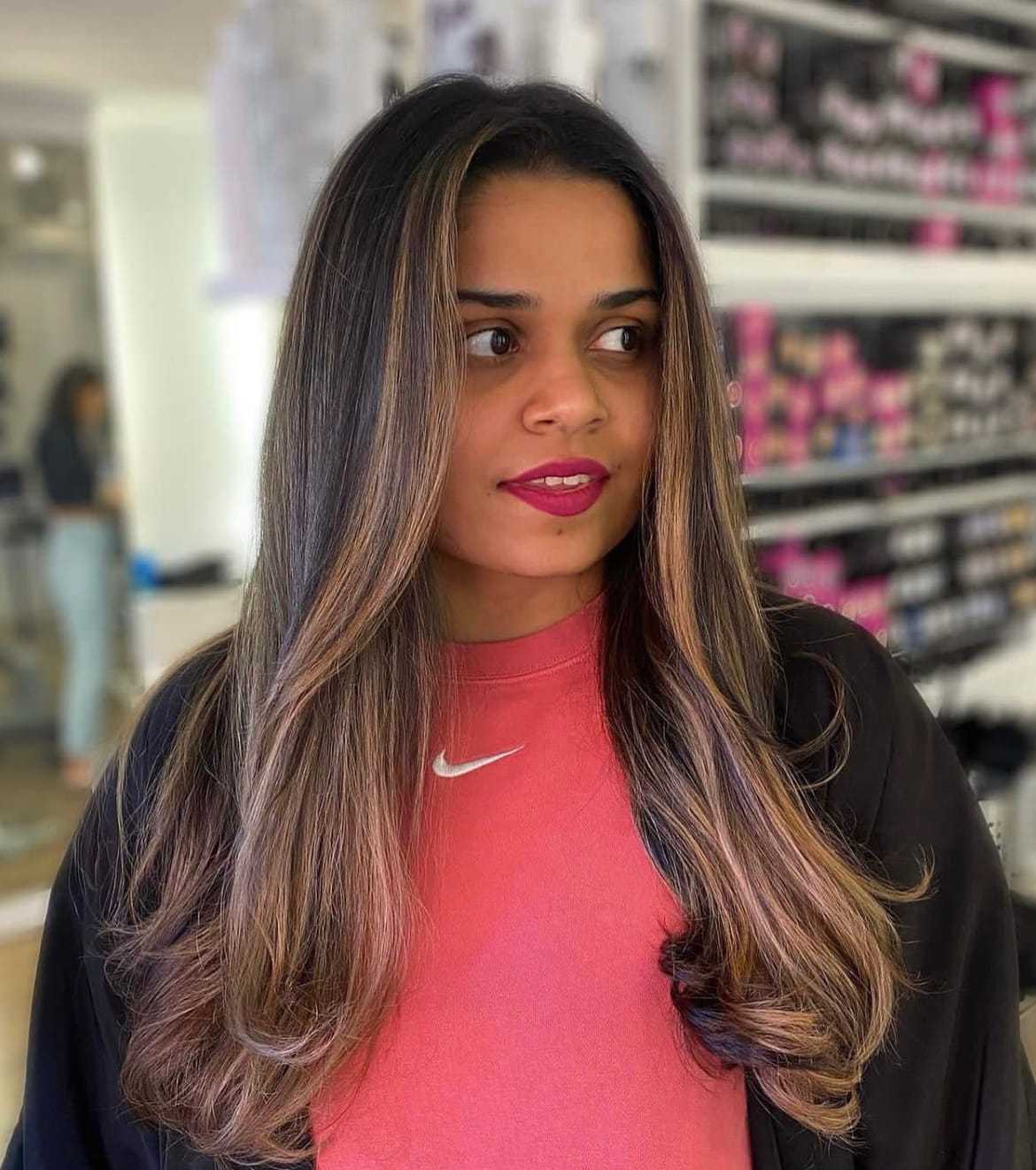 Young woman in a salon with long, styled hair, wearing a red shirt with a logo.