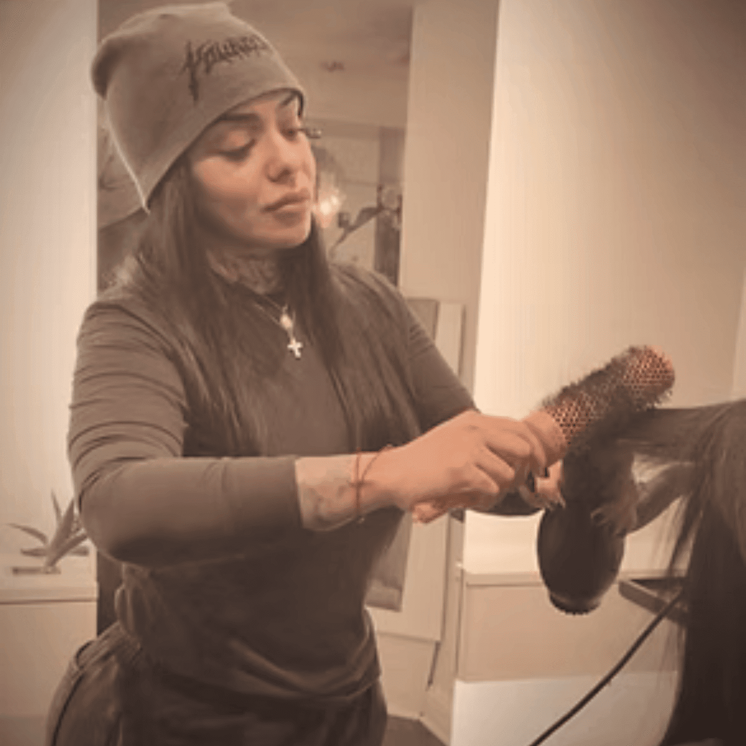 Hairstylist wearing a beanie, using a round brush to style someone's hair in a salon.