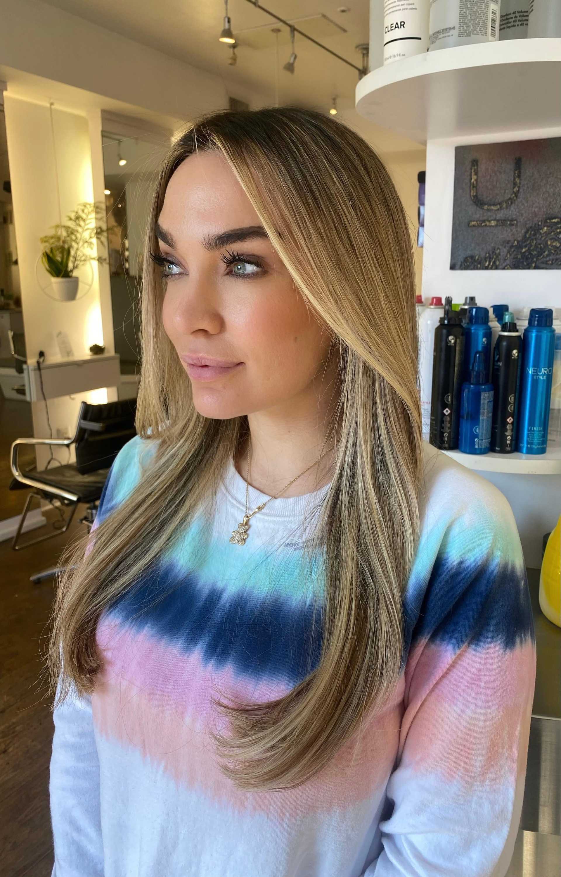 Young woman with long, layered hair at a salon, wearing a colorful tie-dye shirt.